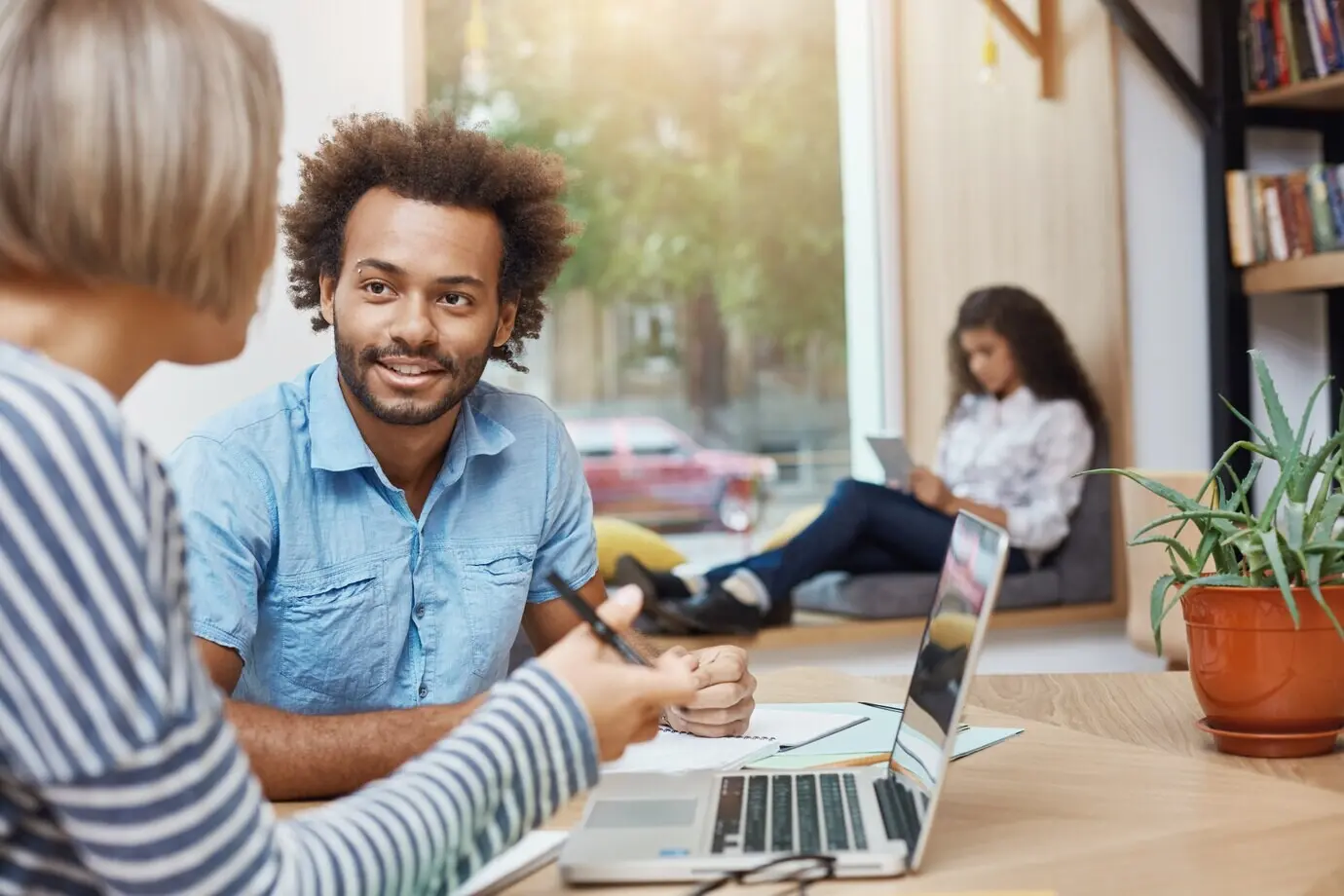 Nahaufnahme eines jungen, gut aussehenden, dunkelhäutigen Freelancers mit lockigem Haar in legerer Kleidung, der in einer Bibliothek sitzt, mit einem Kunden spricht und auf seinem Laptop Beispiele seiner Arbeit zeigt.